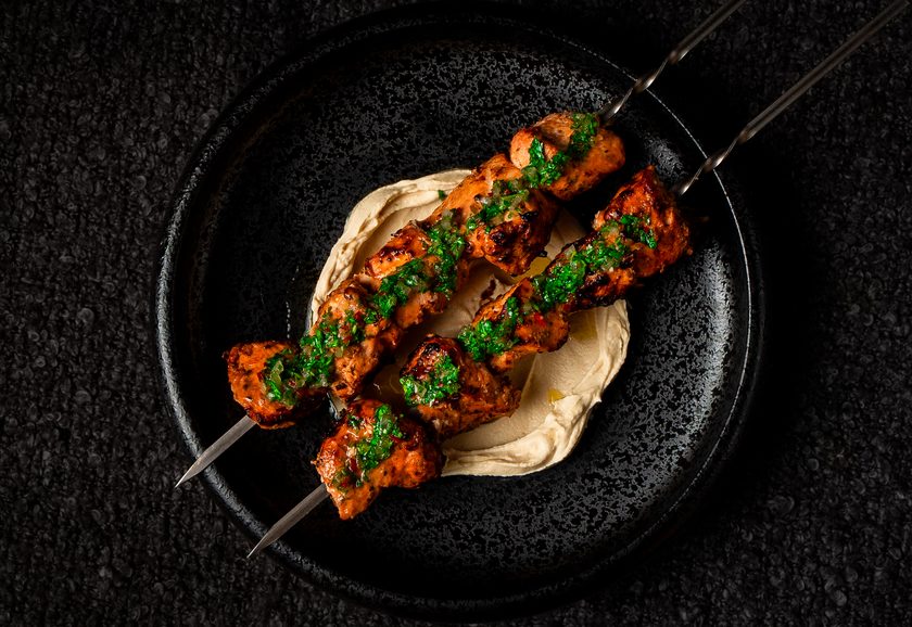 Two charcoal-grilled chicken skewers over hummus with herb sauce