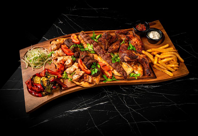 A wooden board of mixed grill with flattened chicken, kebabs, grilled peppers and fries