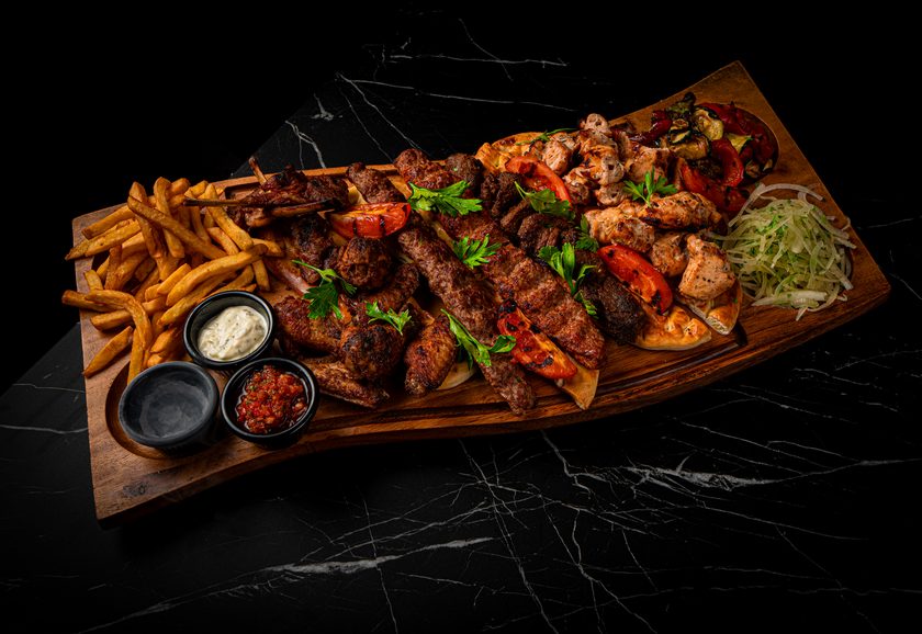 A long wooden board of mixed grill with lamb ribs, kebabs, wings and fries