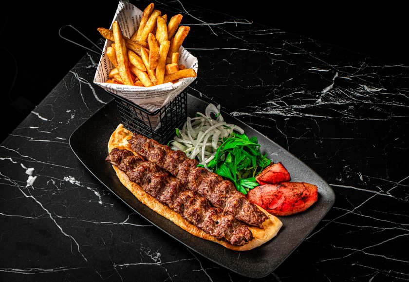 A long minced-meat kebab on flatbread with onions, grilled tomato and pepper, and a basket of fries